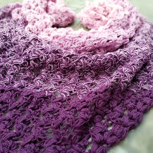 May include: A crocheted shawl with a gradient of pink and purple colors. The shawl is made with a delicate stitch pattern and has a soft, flowing texture.