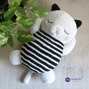 Puede incluir: Un gato de crochet blanco con rayas negras, orejas negras y mejillas rosas. El gato está acostado de lado con los ojos cerrados, como si estuviera durmiendo.