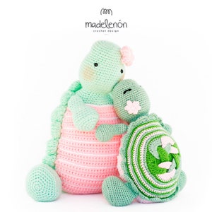 Puede incluir: Dos peluches de tortuga hechos a mano con ganchillo. Uno es verde menta y rosa, el otro es verde y blanco. La tortuga más grande tiene una flor rosa en la cabeza. La tortuga más pequeña tiene una flor en su caparazón. El texto "madelenón crochet design" está en la parte superior.