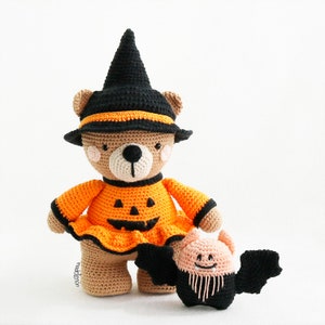 May include: A brown crocheted bear wearing a black witch hat and an orange pumpkin costume. The bear is holding a small crocheted bat with black wings and a pink face.