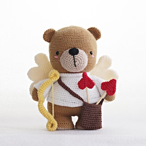 Puede incluir: Un oso de peluche de crochet marrón con alas, que lleva una camisa blanca y lleva un carcaj de flechas con puntas de corazón rojas. El oso sostiene un arco y una flecha amarillos.