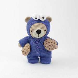 Puede incluir: Un oso de peluche hecho a mano con ganchillo, que lleva un mono azul con capucha y grandes ojos blancos. El oso sostiene dos galletas de ganchillo marrones. La cara del oso es de color canela con una nariz negra y una boca rosada.