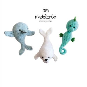 May include: Three crocheted stuffed animals: a light blue dolphin, a white seal, and a light green seahorse. The animals are arranged in a row on a white background. The text "madelenón crochet design" is printed below the animals.