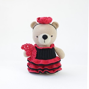 Puede incluir: Oso de peluche de ganchillo con un vestido de flamenco negro y rojo, una flor roja en el pelo y un abanico rojo. El oso tiene un cuerpo color canela, ojos negros y una nariz negra. El vestido tiene volantes.