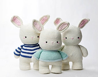 Puede incluir: Tres peluches de conejo blanco de crochet con orejas rosas. Un conejo lleva una camisa a rayas azul y blanca, otro lleva una camisa azul claro y el tercer conejo es liso.