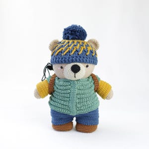 Può includere: Un orsacchiotto all'uncinetto con un cappello blu e giallo con un pompon, un gilet verde, pantaloncini blu e uno zaino marrone. L'orso ha braccia e gambe beige, un naso nero e indossa le cuffie.