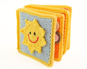 Op de afbeelding: Handgemaakt gehaakt babyboek met een gele zonapplicatie op een blauw vierkant. Het boek heeft gele randen en oranje en roze pagina's. De zon heeft een lachend gezicht met een rode mond en zwarte ogen. Voor baby's en peuters.