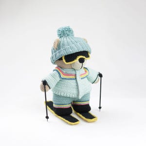 Puede incluir: Un oso de peluche tejido a ganchillo con un atuendo de esquí azul claro, que incluye gorro, chaqueta, pantalones y esquís. El oso lleva gafas de sol negras y bastones de esquí. Los esquís son negros con bordes amarillos.