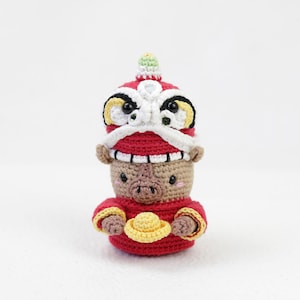 Puede incluir: Figura de cerdo de ganchillo con un disfraz de danza del león rojo con una cara blanca y negra. El cerdo sostiene una moneda de oro. La figura está hecha de hilo y tiene una apariencia linda y caricaturesca.