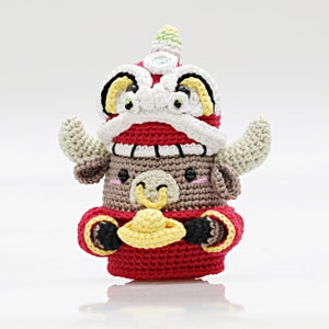Puede incluir: Un juguete de peluche de crochet de un buey marrón que lleva un disfraz de danza del león rojo y blanco. El buey sostiene una moneda de oro en sus patas delanteras.