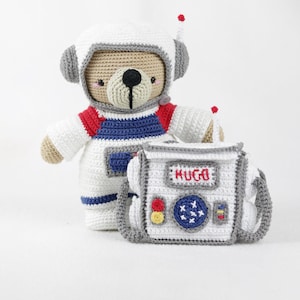 Może przedstawiać: Szydełkowy miś w stroju astronauty, w białym kasku z szarymi akcentami i pasującym kombinezonie kosmicznym. Miś trzyma białą i szarą szydełkową torbę z napisem "HUGO". Torba ma czerwone, żółte i niebieskie detale.