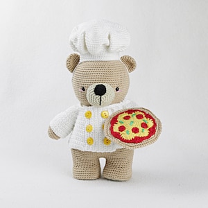 Op de afbeelding: Een gehaakte teddybeer met een witte koksmuts op en een gehaakte pizza in zijn hand. De beer heeft bruine vacht en een zwarte neus.