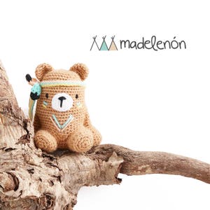 Könnte beinhalten: Ein gehäkelter brauner Bär, der ein türkisfarbenes und weißes Stirnband mit einer Feder trägt. Der Bär sitzt auf einem Stück Treibholz. Der Text "madeleñón" ist im Hintergrund.