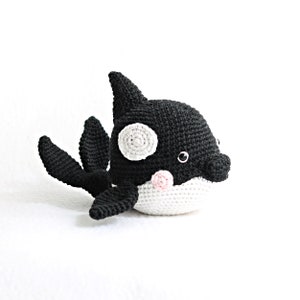 Puede incluir: Un orca de peluche de crochet negro y blanco con una nariz rosa y ojos negros. La orca tiene una mancha blanca en la espalda y una barriga blanca.