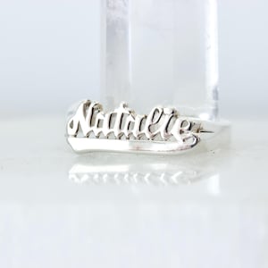 May include: A silver ring with the name "Natalie" in a cursive font. The ring is set against a white background with a clear, cylindrical object behind it. The ring is a piece of jewelry.