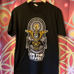 May include: Black t-shirt with a graphic of a samurai helmet in yellow and white. The text "ON THE LEVEL" is printed below the helmet design. The shirt is hanging on a wooden hanger.