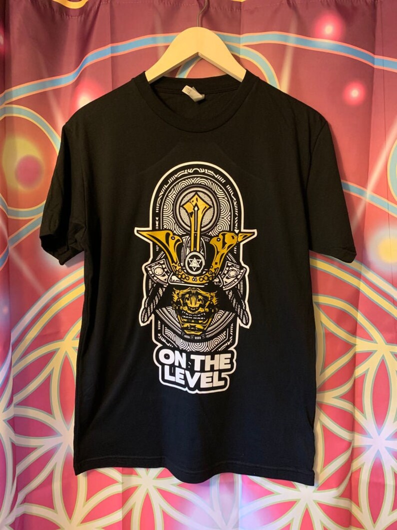 May include: Black t-shirt with a graphic of a samurai helmet in yellow and white. The text "ON THE LEVEL" is printed below the helmet design. The shirt is hanging on a wooden hanger.