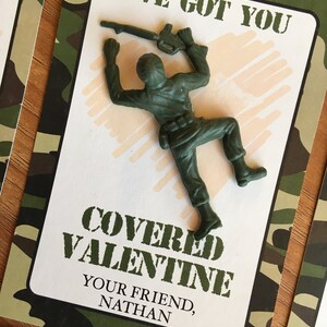 K&ouml;nnte beinhalten: Eine Valentinstagskarte mit einem gr&uuml;nen Spielzeugsoldaten aus Kunststoff. Die Karte hat einen Tarnmuster-Rand und den Text "I've Got You Covered Valentine" und "Your Friend, Nathan."