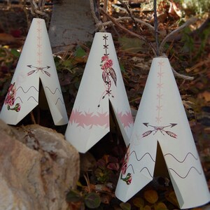 May include: Three cream-colored teepee-shaped decorations with pink and floral designs. Each teepee has a rope for hanging. The decorations are set outdoors among leaves and a rock.