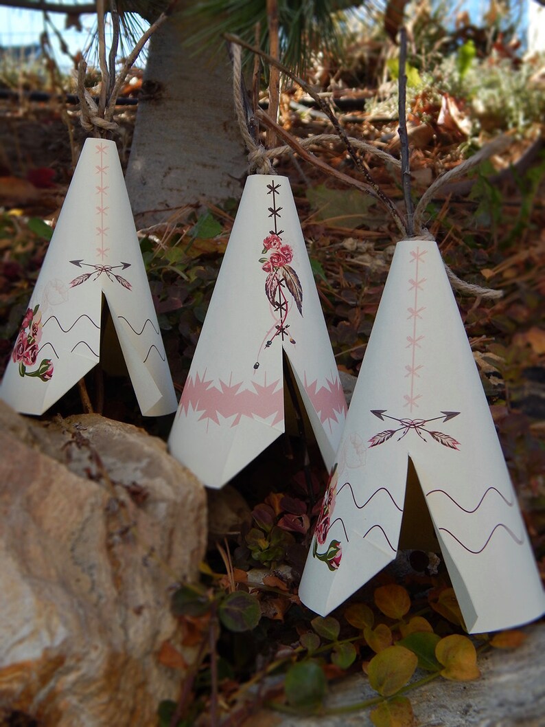 May include: Three cream-colored teepee-shaped decorations with pink and floral designs. Each teepee has a rope for hanging. The decorations are set outdoors among leaves and a rock.