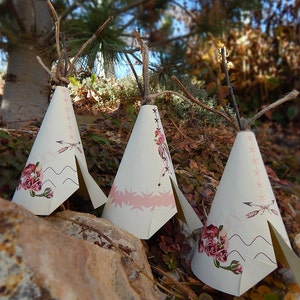 May include: Three white teepee-shaped paper decorations with floral and arrow designs. Each teepee has a brown twig and twine at the top. The teepees are decorated with pink and burgundy floral patterns and sit on a rock.
