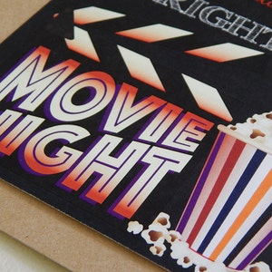 May include: A black card with the words "MOVIE NIGHT" in white, orange, and purple lettering. A popcorn bucket with red, white, blue, and orange stripes is on the right. A film clapperboard is at the top.