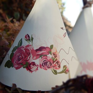 May include: Two white teepee-shaped decorations. One features a large floral design with pink and red roses and green leaves. The other has a pink starburst pattern. Both have a rustic, bohemian aesthetic.