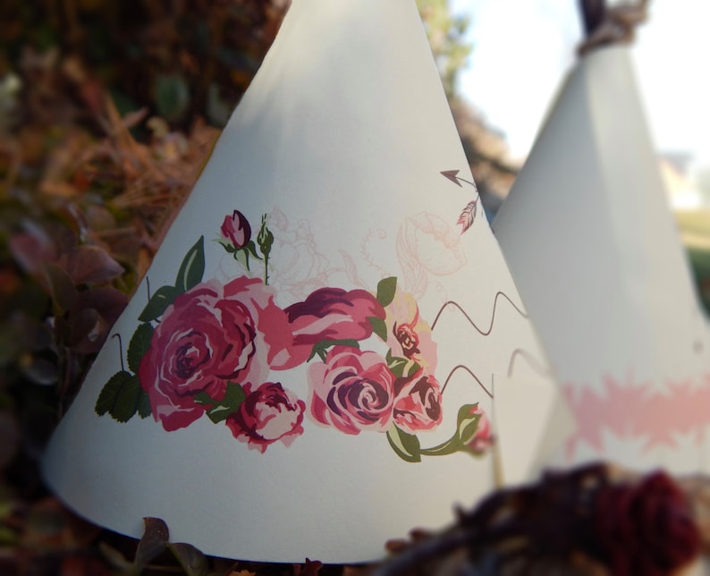 May include: Two white teepee-shaped decorations. One features a large floral design with pink and red roses and green leaves. The other has a pink starburst pattern. Both have a rustic, bohemian aesthetic.