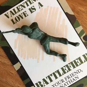 K&ouml;nnte beinhalten: Ein gr&uuml;ner Plastik-Spielzeugsoldat auf einer Valentinstagskarte mit Tarnmuster-Rand. Die Karte lautet "VALENTINE, LOVE IS A BATTLEFIELD" und "YOUR FRIEND, NATHAN". Der Hintergrund ist hellbeige.