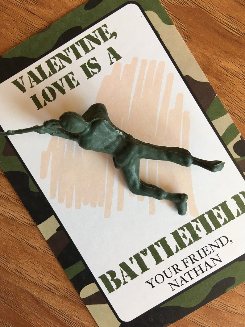K&ouml;nnte beinhalten: Ein gr&uuml;ner Plastik-Spielzeugsoldat auf einer Valentinstagskarte mit Tarnmuster-Rand. Die Karte lautet "VALENTINE, LOVE IS A BATTLEFIELD" und "YOUR FRIEND, NATHAN". Der Hintergrund ist hellbeige.