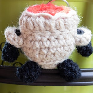 May include: A crocheted cauldron with black arms and legs. The cauldron is filled with red yarn.