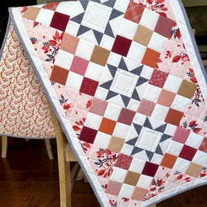 May include: A handmade quilt with a star pattern and a patchwork design. The quilt features squares in shades of red, pink, orange, and brown, with white and gray star accents. Floral patterned fabric borders the quilt.