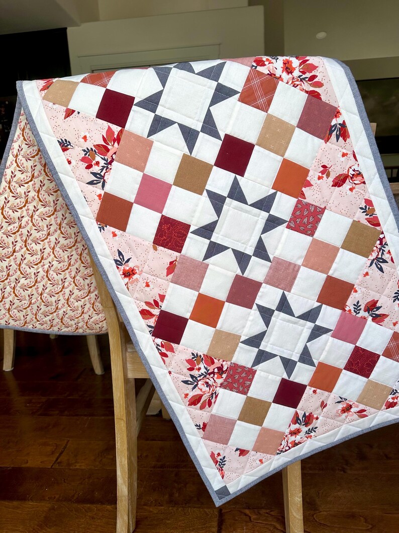 May include: A handmade quilt with a star pattern and a patchwork design. The quilt features squares in shades of red, pink, orange, and brown, with white and gray star accents. Floral patterned fabric borders the quilt.