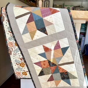 May include: A rectangular quilted table runner featuring two star-shaped blocks made from various patterned fabrics in shades of blue, rust, cream, and brown. The runner is bordered by a gray gingham fabric and a floral print with dark edges.