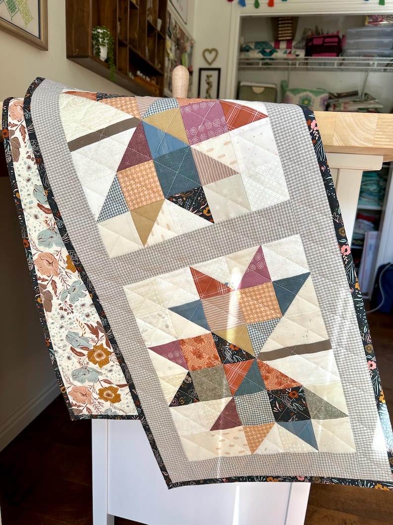 May include: A rectangular quilted table runner featuring two star-shaped blocks made from various patterned fabrics in shades of blue, rust, cream, and brown. The runner is bordered by a gray gingham fabric and a floral print with dark edges.