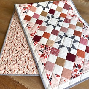 May include: A handmade quilted table runner with a patchwork design. The center features a star pattern with squares in shades of red, pink, white, and brown. The reverse side has a floral pattern on a cream background. The runner is bordered with a gray fabric.