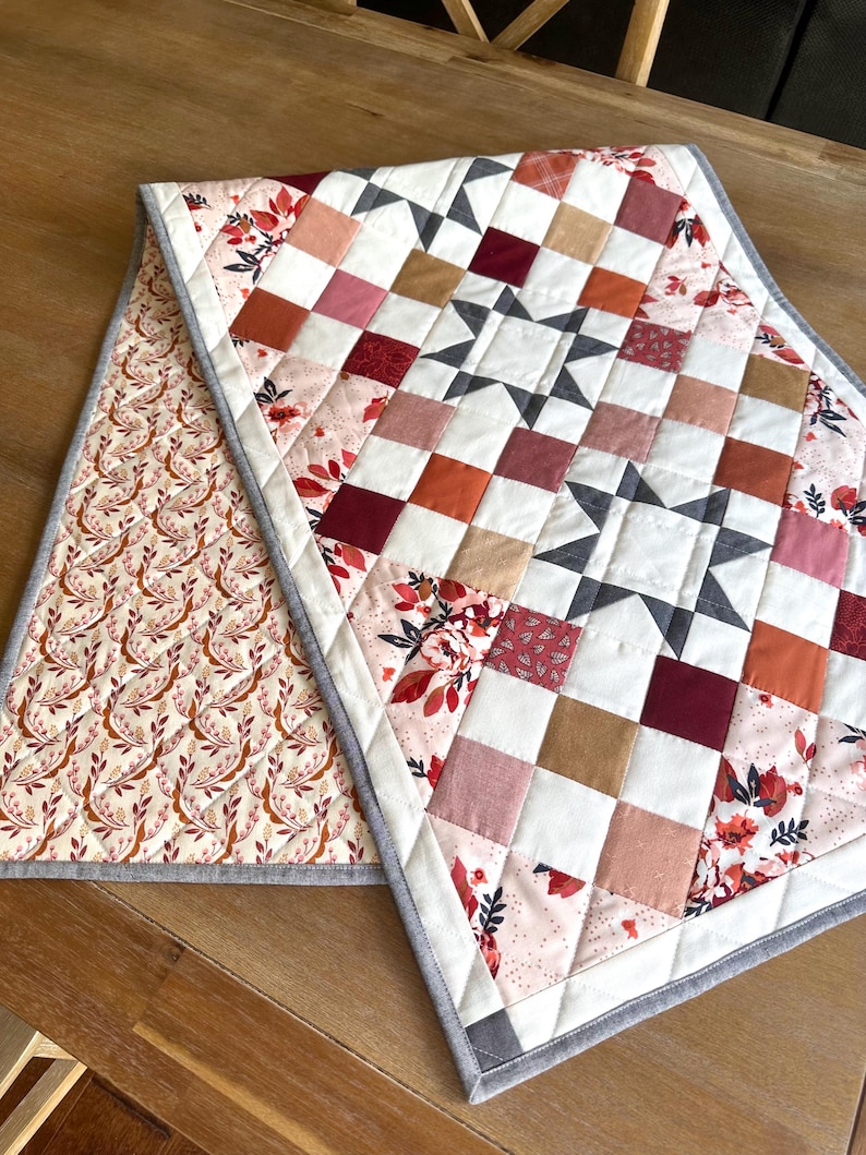May include: A handmade quilted table runner with a patchwork design. The center features a star pattern with squares in shades of red, pink, white, and brown. The reverse side has a floral pattern on a cream background. The runner is bordered with a gray fabric.