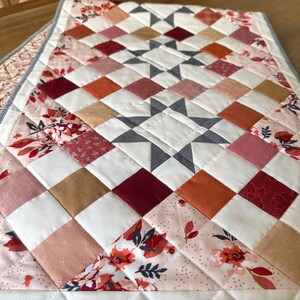 May include: A quilted table runner with a star pattern in the center. The runner features a patchwork design with squares in shades of white, pink, orange, and burgundy. Floral fabric accents the edges, and the runner is bordered with a gray fabric.