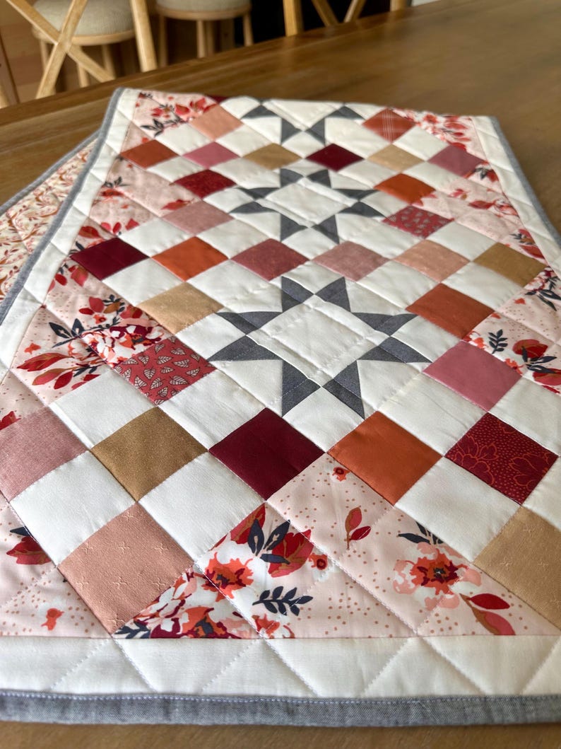 May include: A quilted table runner with a star pattern in the center. The runner features a patchwork design with squares in shades of white, pink, orange, and burgundy. Floral fabric accents the edges, and the runner is bordered with a gray fabric.