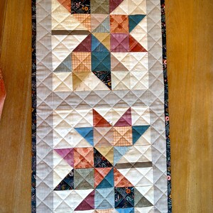 May include: A rectangular quilted table runner featuring two large star patterns composed of various fabric squares in shades of blue, orange, red, and cream. The quilt has a light gray checkered border and a dark floral fabric edge trim.