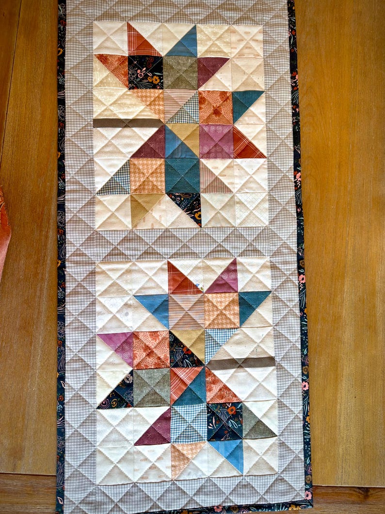 May include: A rectangular quilted table runner featuring two large star patterns composed of various fabric squares in shades of blue, orange, red, and cream. The quilt has a light gray checkered border and a dark floral fabric edge trim.