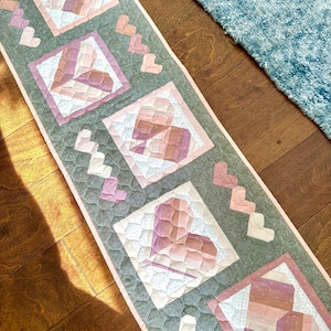 May include: A pink and green quilted table runner with a pattern of hearts. The runner is made of fabric squares sewn together to create a patchwork design.