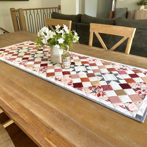 May include: A quilted table runner with a patchwork design in shades of red, pink, and beige, accented with floral patterns and geometric shapes. The runner is placed on a wooden table, with a vase of white flowers and a small jar.