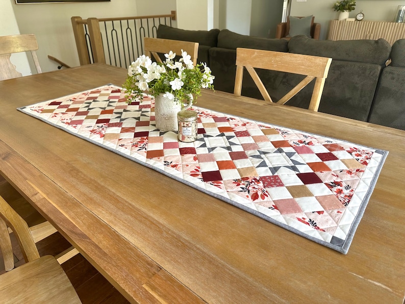 May include: A quilted table runner with a patchwork design in shades of red, pink, and beige, accented with floral patterns and geometric shapes. The runner is placed on a wooden table, with a vase of white flowers and a small jar.