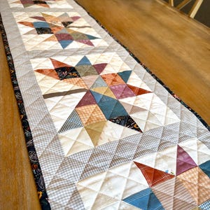 May include: A rectangular quilted table runner featuring a repeating star pattern made from various fabric scraps in shades of rust, blue, cream, and grey. The runner has a dark floral border and is stitched with a diamond-shaped grid pattern.