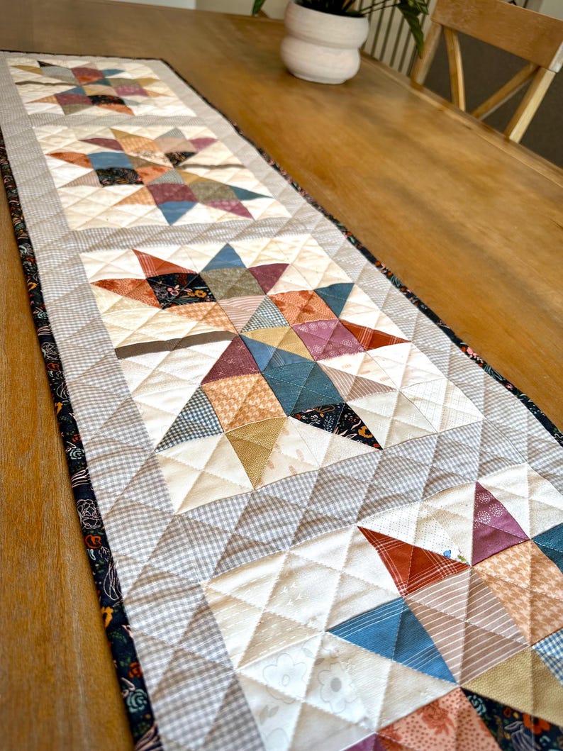 May include: A rectangular quilted table runner featuring a repeating star pattern made from various fabric scraps in shades of rust, blue, cream, and grey. The runner has a dark floral border and is stitched with a diamond-shaped grid pattern.