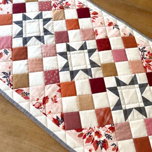 May include: A quilted table runner featuring a patchwork design with squares in shades of white, pink, red, and brown. The runner includes three gray star patterns and floral accents, with a gray border.