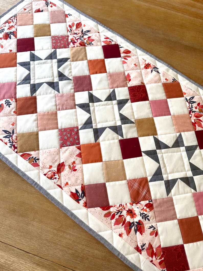 May include: A quilted table runner featuring a patchwork design with squares in shades of white, pink, red, and brown. The runner includes three gray star patterns and floral accents, with a gray border.