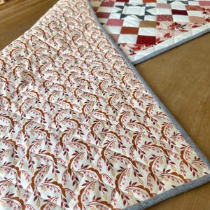 May include: Two quilted placemats with floral and geometric patterns. One features a repeating pattern of brown and pink leaves and berries on a cream background. The other has a patchwork design in shades of red, pink, and brown.