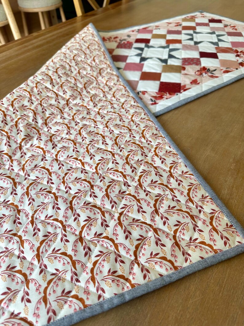 May include: Two quilted placemats with floral and geometric patterns. One features a repeating pattern of brown and pink leaves and berries on a cream background. The other has a patchwork design in shades of red, pink, and brown.